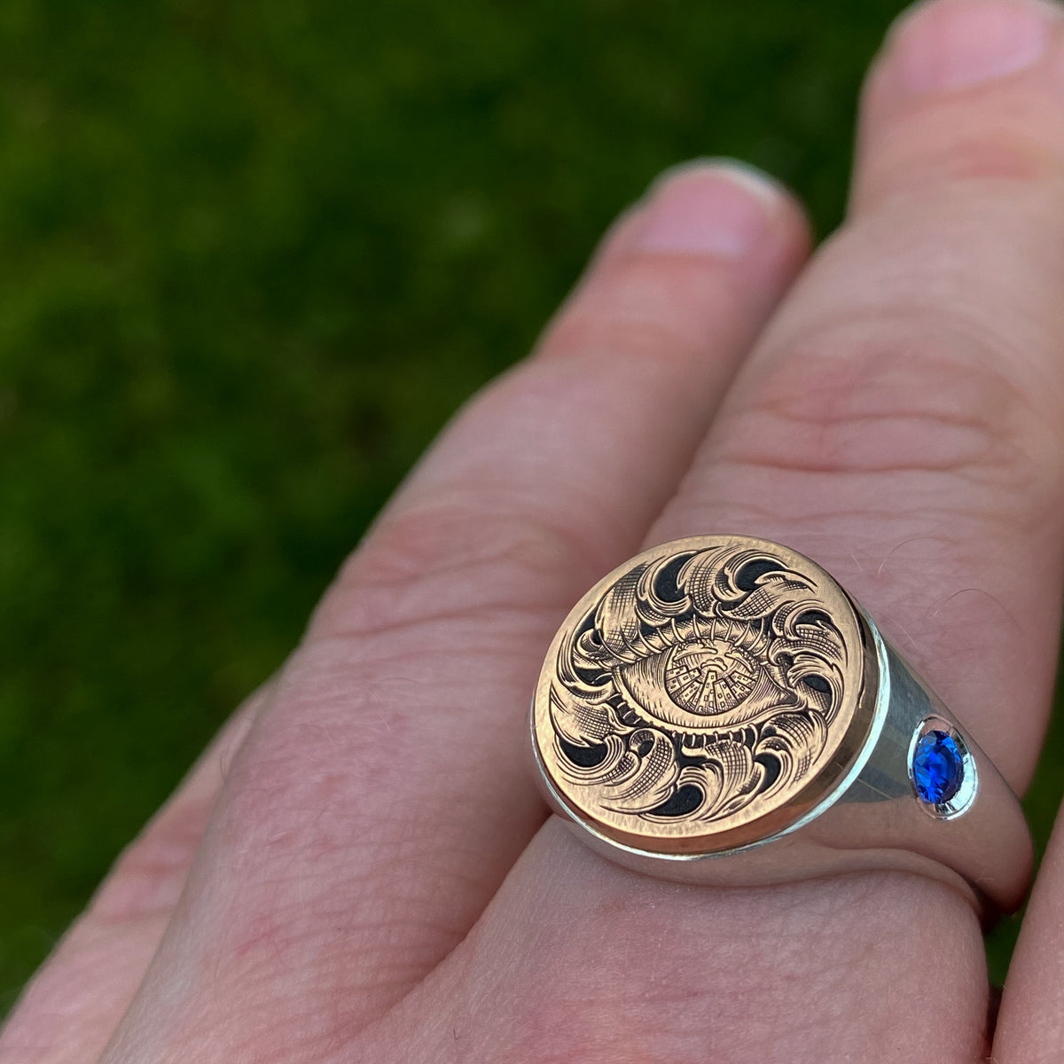 Hand engraved jewellery round signet ring with fantasy leaf scroll work around an eye with city scape in iris. Shoulder set with blue sapphires. Made in 9 carat rose gold and silver. On finger for scale.