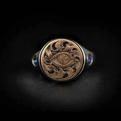 Hand engraved jewellery round signet ring with fantasy leaf scroll work around an eye with city scape in iris. Shoulder set with blue sapphires. Made in 9 carat rose gold and silver. On black background face on.