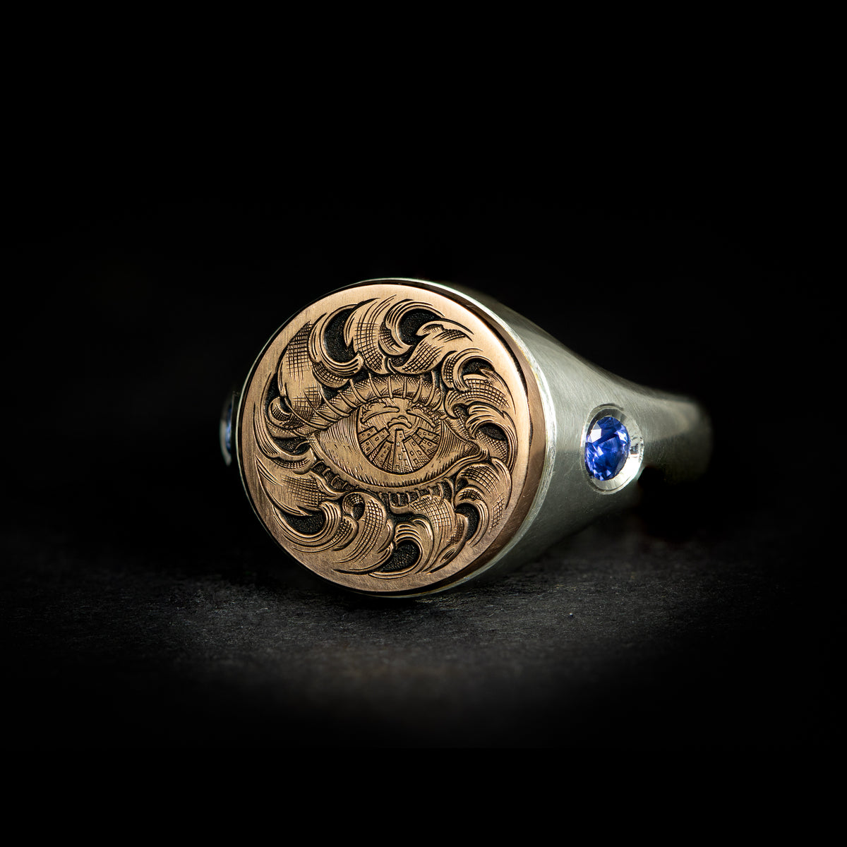 Hand engraved jewellery round signet ring with fantasy leaf scroll work around an eye with city scape in iris. Shoulder set with blue sapphires. Made in 9 carat rose gold and silver. On black background angled shot.