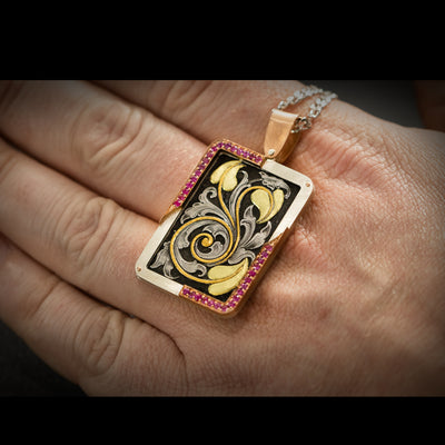 Jewellery pendant hand engraved featuring deep relief scrolls with inlaid fine gold and green gold. Has a frame of silver, rose gold and pin sapphires. Lying against hand for scale