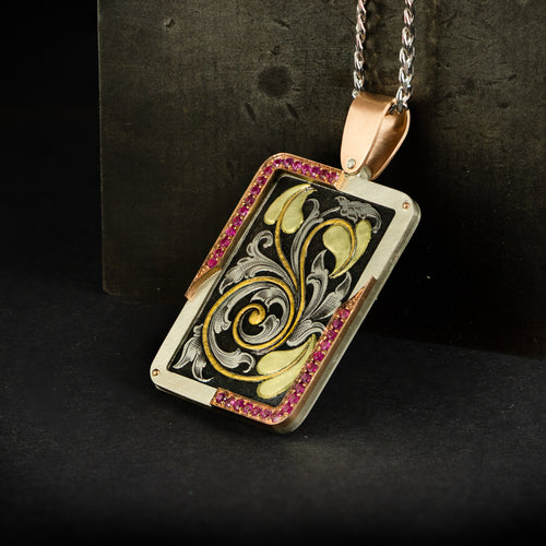 Jewellery pendant hand engraved featuring deep relief scrolls with inlaid fine gold and green gold. Has a frame of silver, rose gold and pin sapphires. Lying against steel back drop