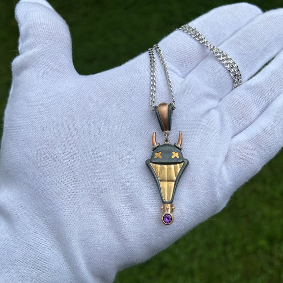 Large Smile Please hand crafted jewellery pendant. Hand crafted smile motif with horns in oxidised silver, 18ct yellow gold, rose gold, white gold and fine gold set with one amethyst. On white gloved hand for scale
