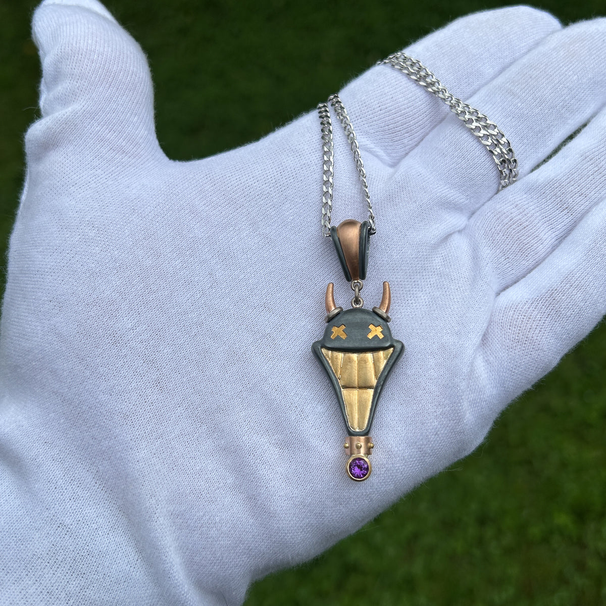 Large Smile Please hand crafted jewellery pendant. Hand crafted smile motif with horns in oxidised silver, 18ct yellow gold, rose gold, white gold and fine gold set with one amethyst. On white gloved hand for scale