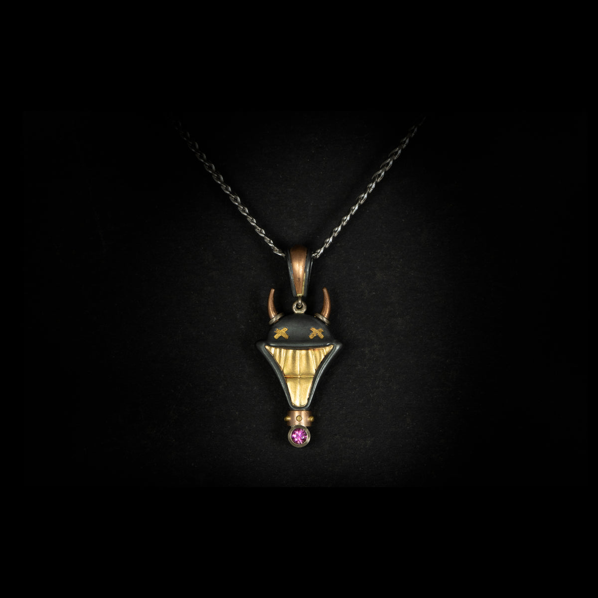 Smile Please hand crafted jewellery pendant. Hand crafted smile motif with horns in oxidised silver, 18ct yellow gold, rose gold, white gold and fine gold set with one round pink sapphire. On chain on black background.