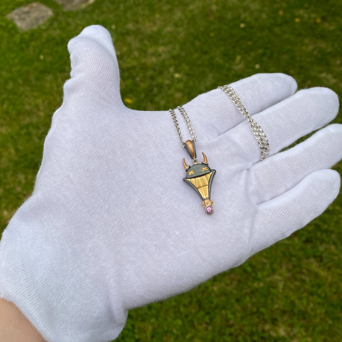 Smile Please hand crafted jewellery pendant. Hand crafted smile motif with horns in oxidised silver, 18ct yellow gold, rose gold, white gold and fine gold set with one round pink sapphire. On chain held in white gloved hand for scale