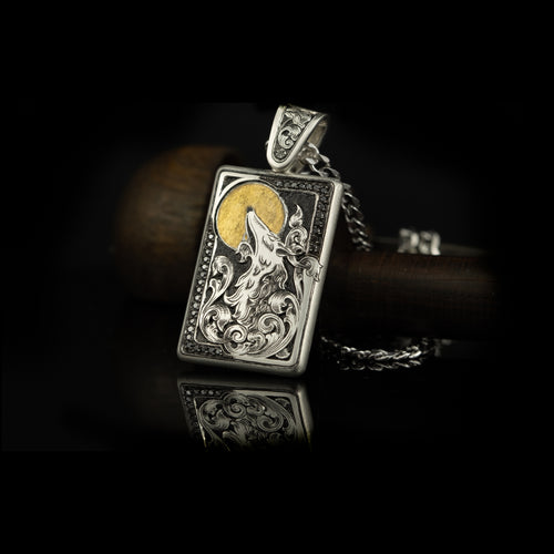 Hand engraved jewellery pendant featuring a fantasy wolf design howling at a 24 carat fine gold moon. Wolf is surrounded by scroll work and set with black diamonds. Made in silver. Displayed on black background.